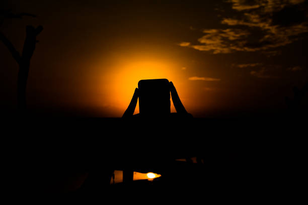A silhouette of hands holding a phone as it frames the setting sun and its reflection in the calm water.