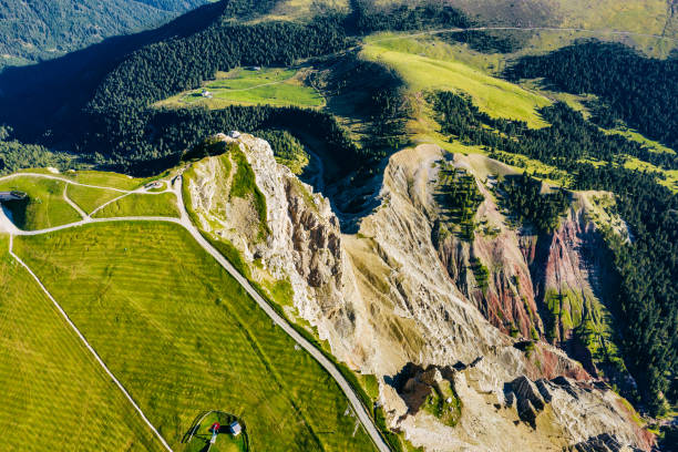 Steep rock walls rise dramatically from the valley floor, their sharp formations contrasting with the soft greens of surrounding meadows. The vivid blue sky in the background.