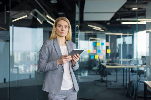 istock Serious and confident businesswoman at workplace standing by window with tablet computer in hands, woman in business suit looking at camera, working inside office 2197444757