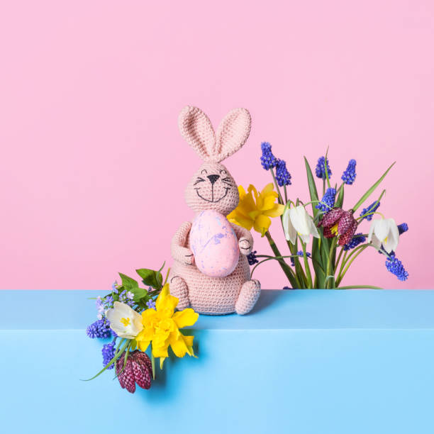 Knitted toy pink bunny with Easter egg and bouquet of spring flowers on blue table and purple background. Concept of spring, Easter, Mother's Day