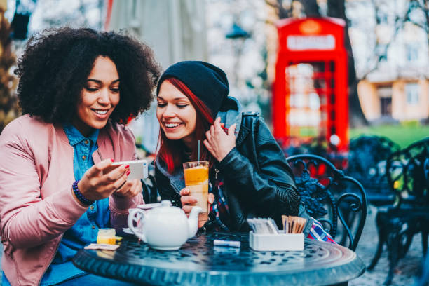 Friends enjoying a cozy outdoor cafe moment Two friends seated at an outdoor cafe, sharing a moment over drinks and a smartphone. Dating girl stock pictures, royalty-free photos & images