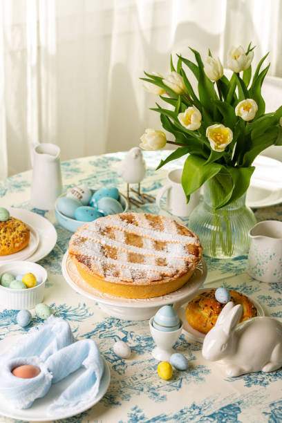istock Interior view of served easter round dining table, Italian Easter cake, shortcrust pie with ricotta cheese, Pastiera Napoletana, colorful eggs, buns, easter bunny and bird sculptures, vase with tulips flowers. Vertical image. 2197458472