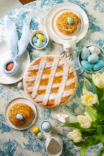 istock Served easter table for brunch, directly above. Italian Easter cake, shortcrust pie with ricotta cheese, Pastiera Napoletana, colorful eggs, buns, easter bunny and bird sculptures, vase with tulips flowers. Blue and white table cloth. Vertical image. 2197458474