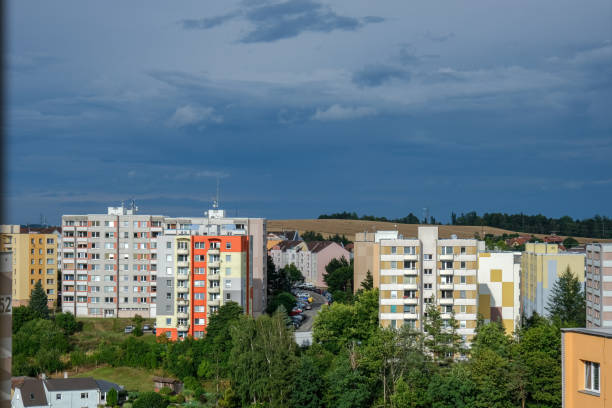 A group of colorful residential apartment buildings surrounded by greenery, with a dramatic stormy sky in the background. The image captures an urban landscape with modern housing.