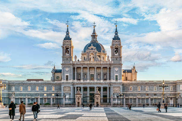Almudena Cathedral in Madrid with its impressive neoclassical facade under a serene blue sky. People walk through the plaza, enjoying the grand architecture and historic charm of the landmark. Madrid, Spain - November 30, 2021