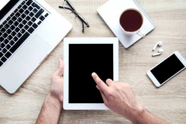Male hands holding tablet computer with laptop, smartphone and cup of tea on table