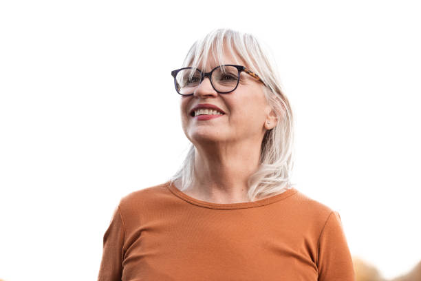 A joyful senior woman with glasses smiles confidently outdoors, wearing a casual orange top. Captures the essence of positive aging and confidence in natural daylight.
