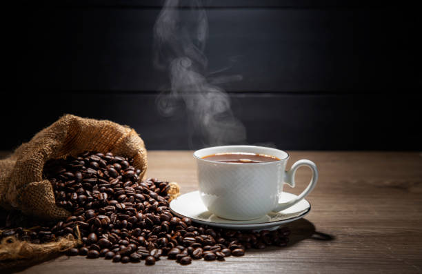 istock Hot coffee in a white coffee cup With roasted coffee beans on a wooden table, dark black background. 2197498342