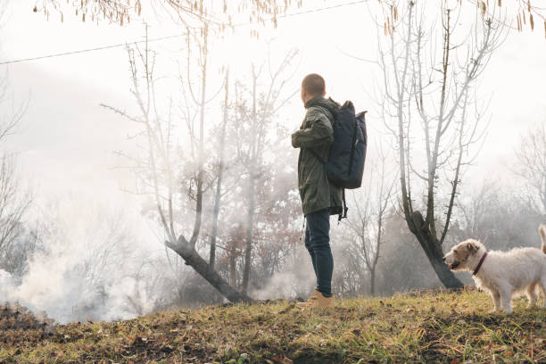A young man with a large hiking backpack walks through the morning mist, holding binoculars in his hand, enjoying the silence of nature and the magic of adventure.