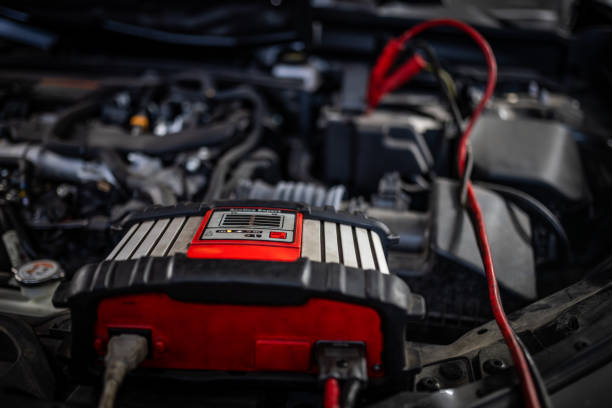 A close-up view of a charging device connected to a car engine in a workshop. The scene features detailed engine components and vibrant red cables, highlighting automotive maintenance processes in a professional setting.