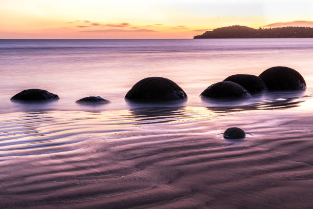 The Moeraki Boulders at sunrise on Koekohe Beach, New Zealand. The smooth, spherical boulders emerge from the tide, partially submerged in the shimmering water, creating a surreal and otherworldly scene.  I used the long exposure to soften the waves, giving the ocean a misty, dreamlike quality, while the golden hues of dawn contrast with the deep blues and purples of the receding night. The composition highlights the intricate details of the sand ripples and reflections, enhancing the timeless and mystical feel of this iconic location.