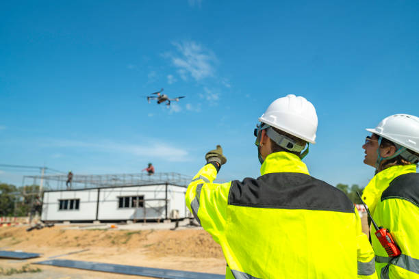 istock Two engineers in high visibility jackets and helmets observing a drone flying over a construction site, with machinery and modular buildings, highlighting technology and site monitoring. 2197510046