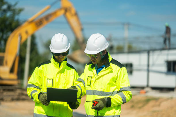istock Two engineers in high-visibility jackets and helmets reviewing plans on a laptop at a construction site with an excavator in the background, representing safety, precision, and technological planning. 2197510206