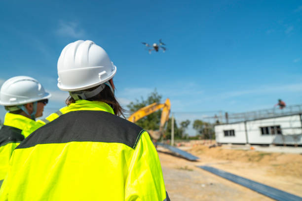 istock Two engineers in high visibility jackets and helmets observing a drone flying over a construction site, with machinery and modular buildings, highlighting technology and site monitoring. 2197511782