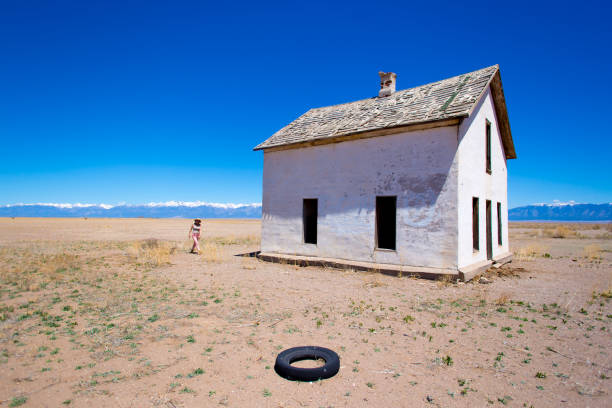 A hauntingly beautiful scene of an abandoned Bandan farmhouse beside a gravel pit in the rolling hills of western South Dakota. The once-vibrant farmhouse now sits in solitude, showing signs of wear and neglect, while the gravel pit nearby adds to the atmosphere of abandonment. The bright blue sky  contrasts with the desolate setting, and the distant I-90 highway cuts through the vast, open landscape. This image evokes a sense of nostalgia and forgotten history, ideal for themes of abandonment, rural decay, and Western landscapes.