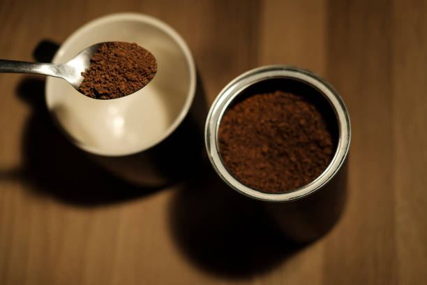 Metal spoon lifting instant coffee granules above an empty mug next to an open tin of instant coffee on a wooden table, ready to be mixed with hot water