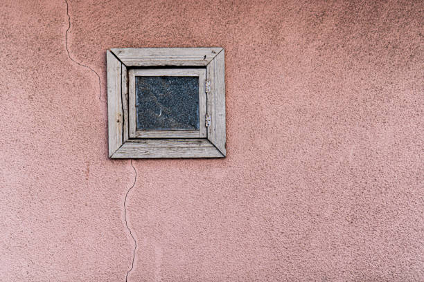A small, weathered wooden window with a cracked glass pane is set into a textured pink stucco wall. The aged frame, with its rough, faded wood, stands out against the soft, muted pink backdrop. A visible crack runs from the window down the wall, emphasizing the effects of time and exposure.