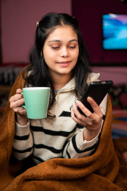 istock Relaxed young woman sitting comfortably in bed with cozy blanket and using a smartphone while drinking a hot cup of coffee in winters and enjoying her leisure time at home. 2197547882