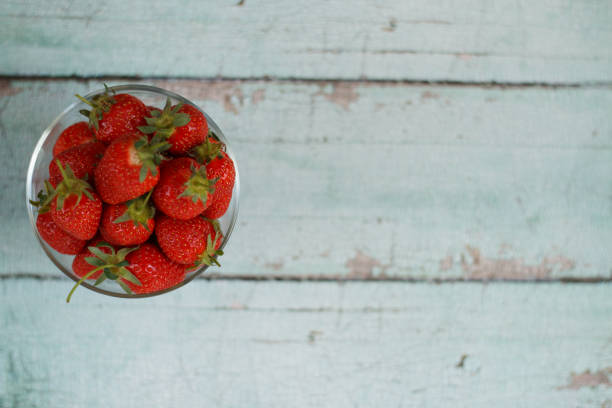 Fresh strawberries in a glass bowl on a rustic wooden table, showcasing their vibrant red color and natural texture. The transparent bowl adds a delicate touch, allowing the rich tones of the fruit to stand out against the rustic background.