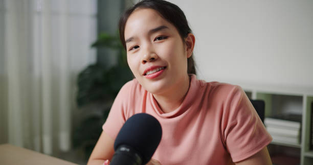 istock A young woman in a light pink shirt is sitting at a table and talking or explaining something with a bright expression in front of the camera. 2197556180