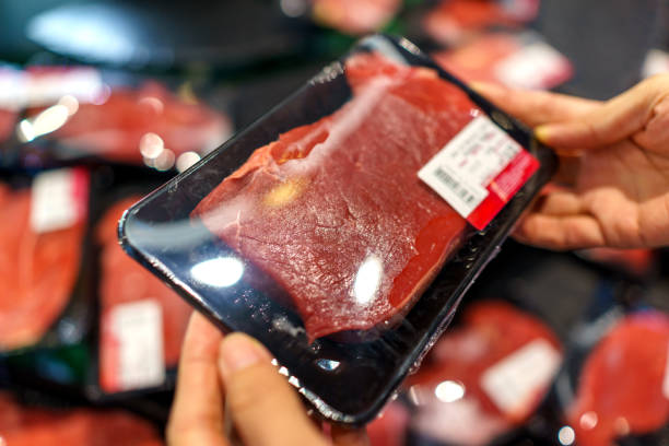 Shopper holding a tray of packaged raw beef while browsing the meat section in a supermarket.