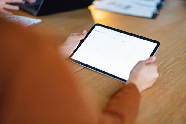 A colleague holds a digital tablet displaying a calendar during an office meeting, showcasing collaboration and technology usage. The focus on digital tools highlights modern workplace strategies and efficiency in planning.