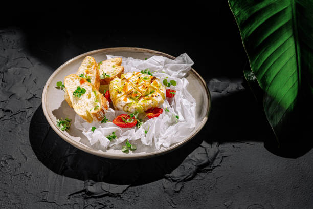A plate showcases a creamy cheese surrounded by sliced tomatoes and fresh bread. The dish is elegantly arranged with herbs on a textured black surface.