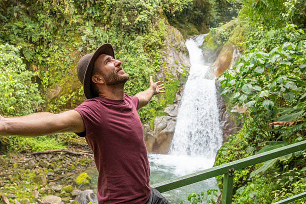 Man in hat stands joyfully with arms open by a vibrant waterfall. Hiker revels in nature's beauty amid lush greenery. Outdoor enthusiast embracing wilderness adventure.