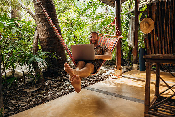 Man lounging in a hammock, working on a laptop amidst lush tropical plants. Relaxed digital nomad lifestyle.