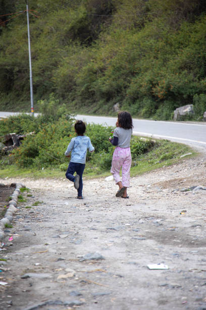 Two children walk on a dirt pathway surrounded by lush greenery beside a quiet roadside. Their casual strides and natural setting evoke a sense of freedom and exploration in a rural environment.