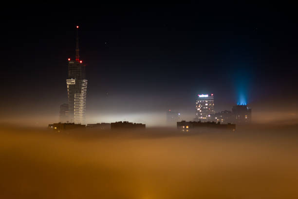 Fotografia przedstawia nowoczesny wieżowiec otoczony mgłą, uchwycony nocą w mieście. Na zdjęciu widoczne są kontrastujące światła budynków oraz delikatny efekt mgły, który dodaje scenie tajemniczości i atmosfery. Idealne dla projektów związanych z architekturą, miejskimi krajobrazami i nocnymi pejzażami.
