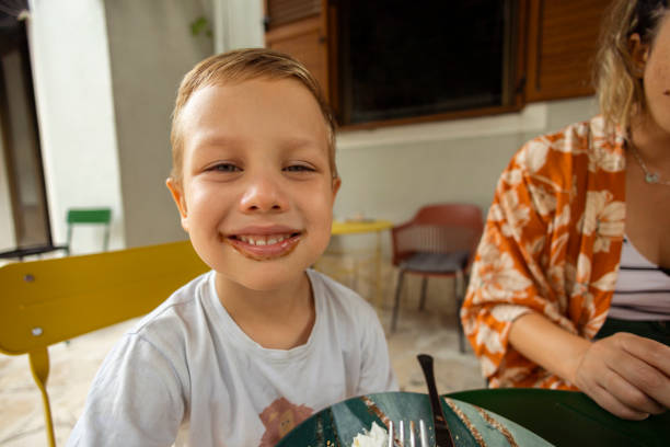 Cute little boy sitting at dinning room table making funny faces and smiling while having breakfast at home