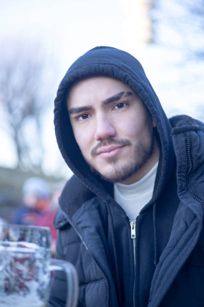 A young man in a black hoodie and jacket sits outdoors with a relaxed expression, enjoying a casual moment. The bright background and natural light emphasize his approachable and calm demeanor