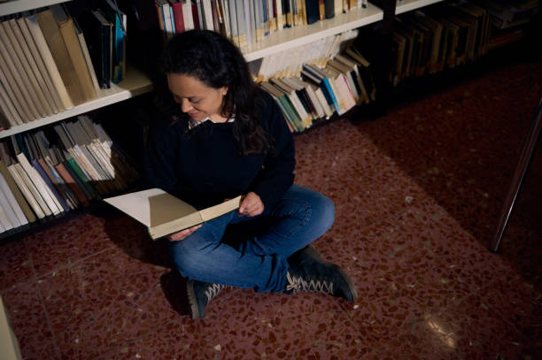 istock Young woman reading a book while sitting on library floor 2197609604