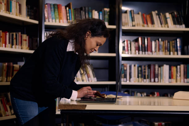 istock Focused woman using laptop at library surrounded by bookshelves 2197609619