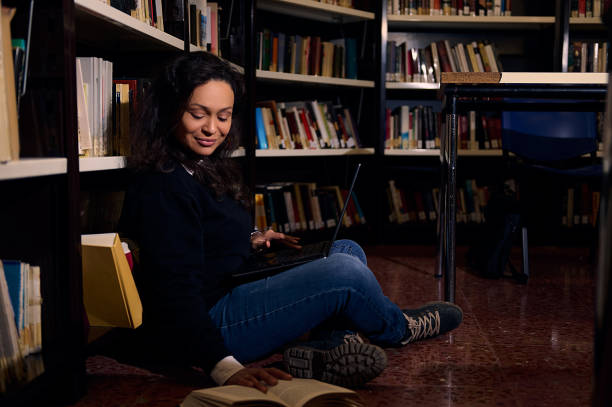 istock Young woman studying with a book and laptop in a cozy library corner 2197609633