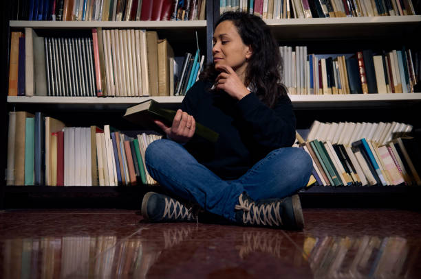 Woman reading book while sitting in front of library shelves
