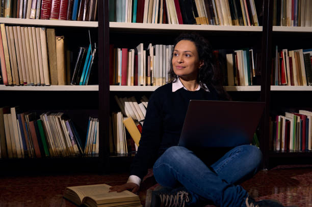 istock Woman sitting in library with laptop and book, smiling thoughtfully 2197609685