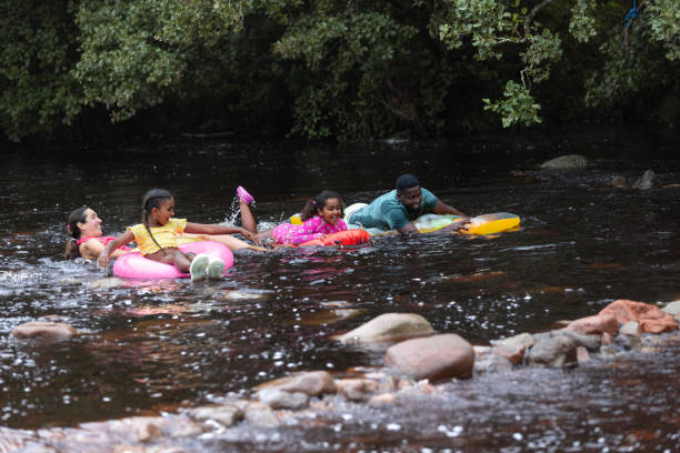 Wide shot of a family in a river in Northumberland, North East of England. They are on a family day out. Everyone is in swimming costumes as they are playing and floating along a river on inflatables. \n\nVideos are available similar to this scenario.