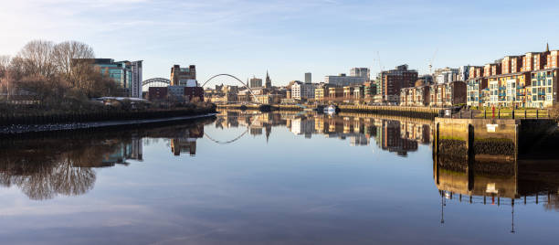 A panoramic view of the River Tyne in Newcastle in the North East of England. It is a still serene scene with the still water reflecting like a mirror the outline of the buildings and bridges along the wharf and quayside with a beautiful calm blue sky.