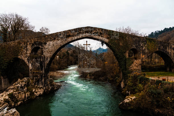 Ancient stone bridge arching over a flowing river, with a cross visible through the central arch. Roman Bridge Cangas de Onis