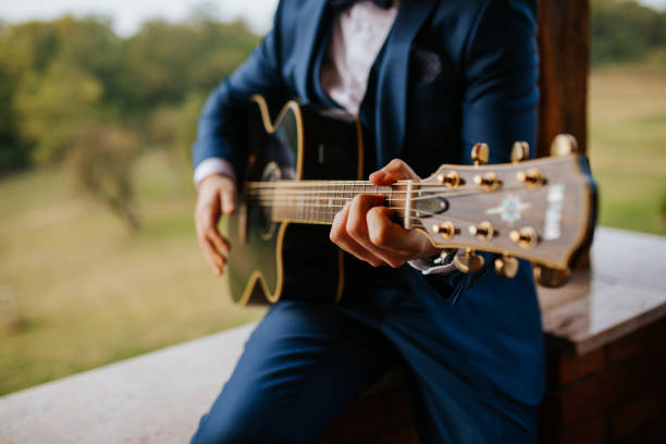 A groom in a stylish navy-blue suit plays an acoustic guitar, capturing a heartfelt and intimate moment. A perfect blend of music, romance, and elegance in a natural outdoor setting.