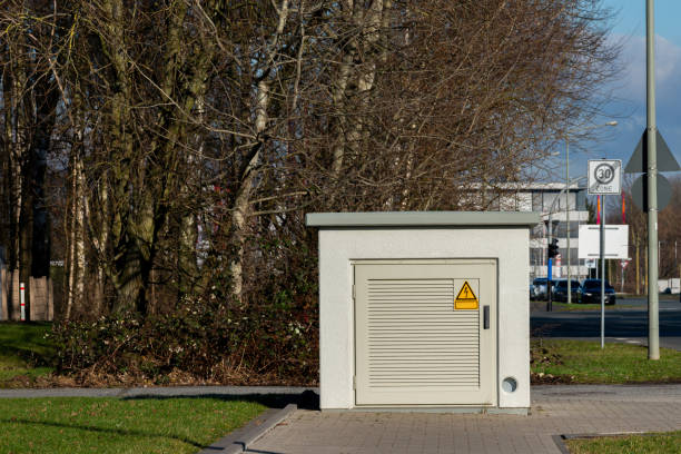 A utility box sits peacefully beside a tree-lined path in an urban area, showcasing its solid construction and safety warning symbol against a backdrop of clear blue skies.