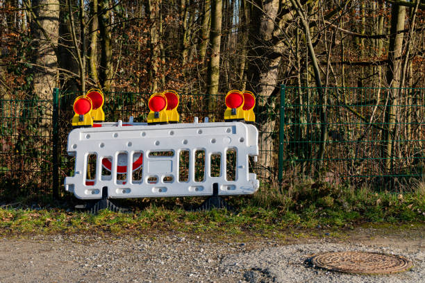 A white construction barrier with bright orange warning lights is positioned beside a gravel path near a wooded area. The surrounding trees are bare, indicating winter, and the sky is clear.