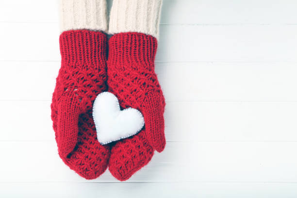 Hands in knitted mittens holding heart on white wooden table