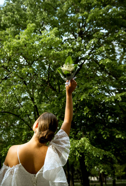 bride with a bouquet in her hand. the bride stands with her back in the frame. holds a bouquet of lilies of the valley in her raised hand. a brunette girl against the background of green trees.