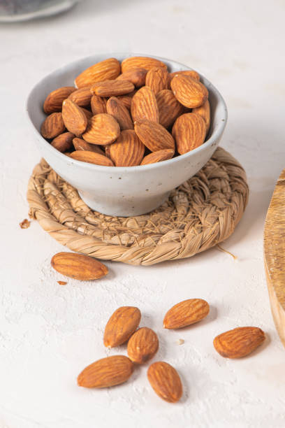 Bowl of almonds on a woven mat, with some almonds scattered on a white surface, showcasing a healthy snack option in a rustic setting