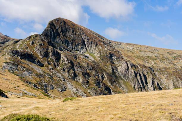 Majestic mountain terrain revealing rugged rock formations, verdant grassy slopes beneath azure sky with wispy cloud patterns, highlighting pristine wilderness