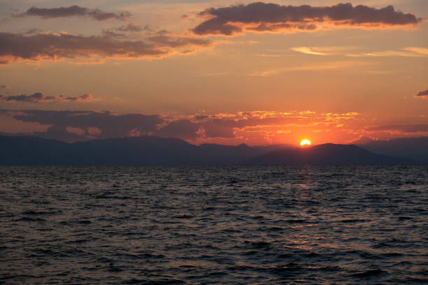 A stunning sunset paints the sky in warm hues of orange and red, casting a golden glow over the rippling sea. The distant silhouette of mountains adds depth to this serene and captivating coastal scene.