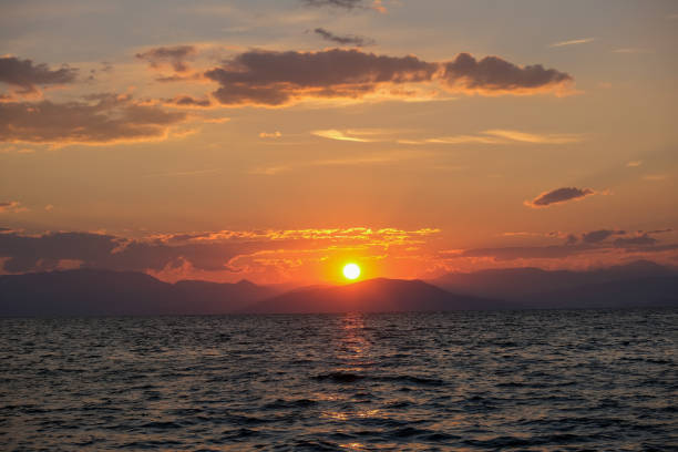 A stunning sunset paints the sky in warm hues of orange and red, casting a golden glow over the rippling sea. The distant silhouette of mountains adds depth to this serene and captivating coastal scene.